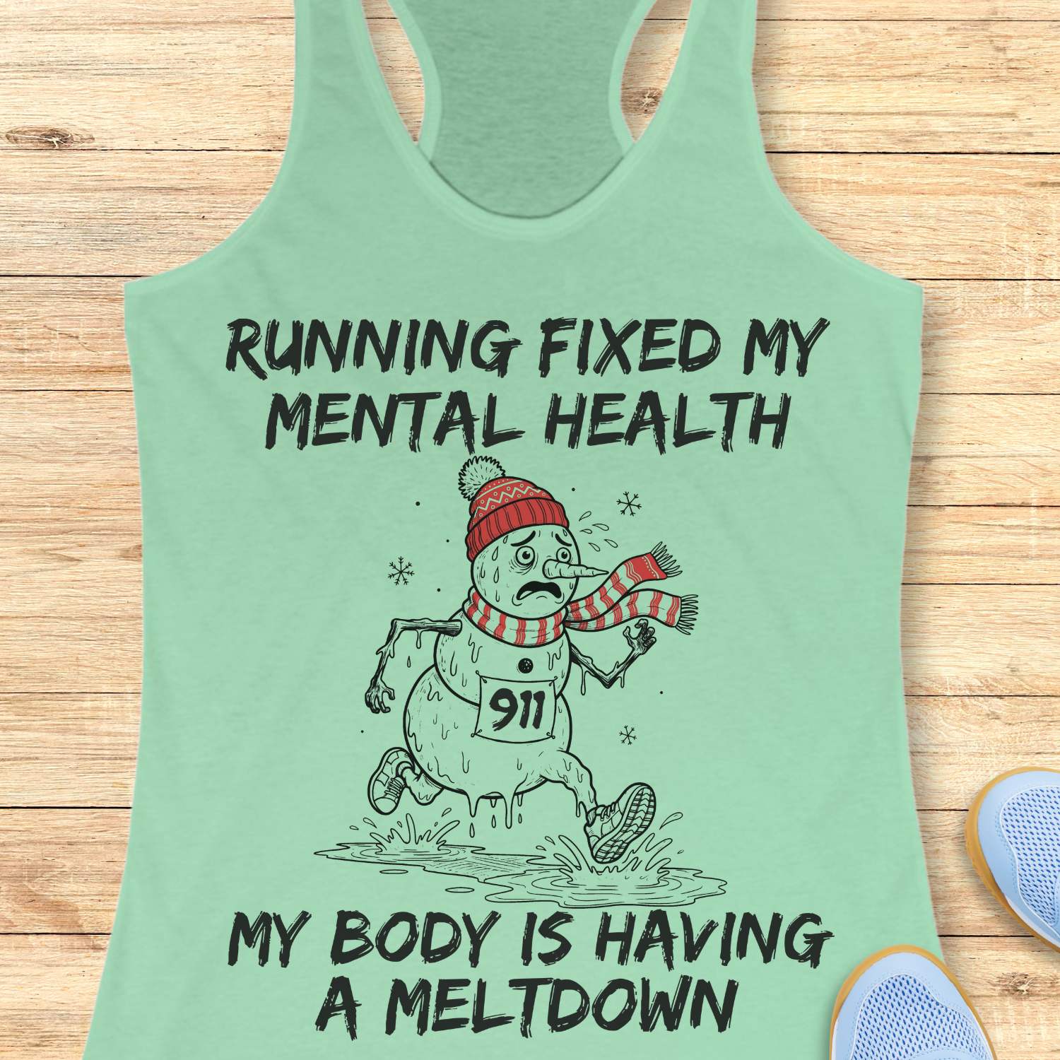 mint racerback tank top displayed on a light wooden surface featuring a humorous running-themed Christmas design. The graphic shows a melting snowman wearing a red knit hat, striped scarf, running shoes, and race bib number “911,” splashing through a puddle while running. Bold black text above and below the illustration reads: “RUNNING FIXED MY MENTAL HEALTH” and “MY BODY IS HAVING A MELTDOWN.” Partial blue athletic shoes are visible in the lower corners of the image.