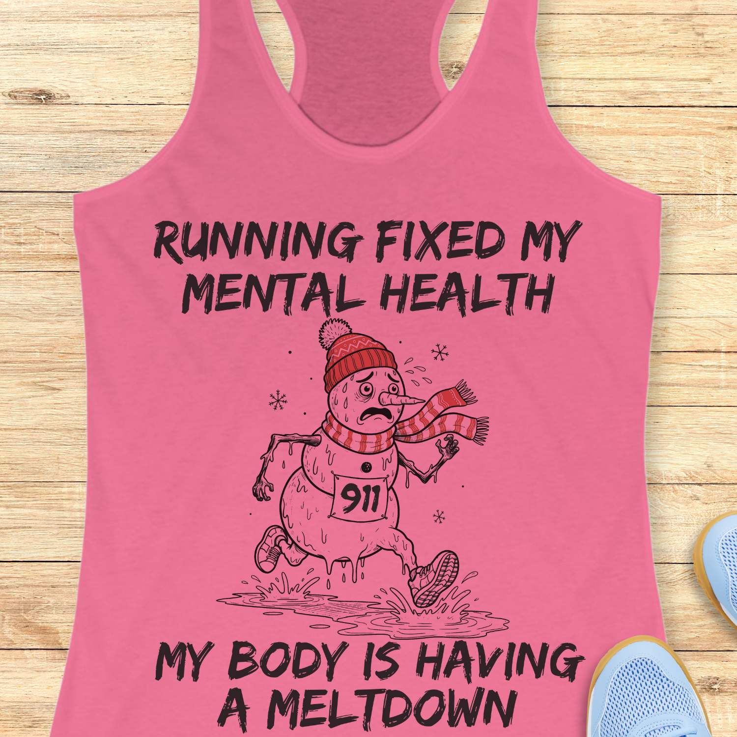 pink racerback tank top displayed on a light wooden surface featuring a humorous running-themed Christmas design. The graphic shows a melting snowman wearing a red knit hat, striped scarf, running shoes, and race bib number “911,” splashing through a puddle while running. Bold black text above and below the illustration reads: “RUNNING FIXED MY MENTAL HEALTH” and “MY BODY IS HAVING A MELTDOWN.” Partial blue athletic shoes are visible in the lower corners of the image.