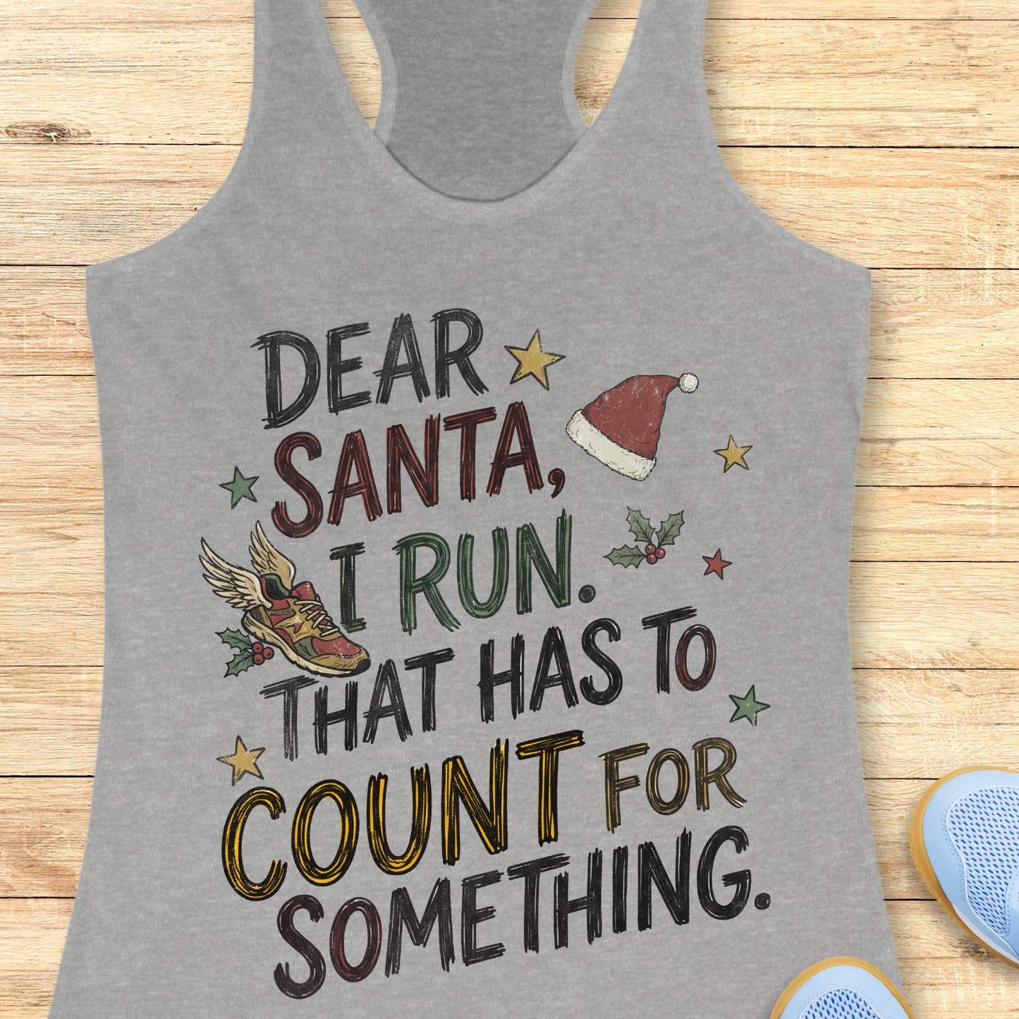 grey racerback tank top displayed on a light wooden surface, featuring a festive running-themed typographic design. The graphic includes the humorous text: “DEAR SANTA, I RUN. THAT HAS TO COUNT FOR SOMETHING.” Decorative holiday elements surround the text, including a red Santa hat, a winged running shoe, holly sprigs with berries, and colorful stars. The word “COUNT” is highlighted in yellow, while the remaining text appears in black. Partial blue athletic shoes are visible in the lower corners, rei