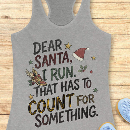 grey racerback tank top displayed on a light wooden surface, featuring a festive running-themed typographic design. The graphic includes the humorous text: “DEAR SANTA, I RUN. THAT HAS TO COUNT FOR SOMETHING.” Decorative holiday elements surround the text, including a red Santa hat, a winged running shoe, holly sprigs with berries, and colorful stars. The word “COUNT” is highlighted in yellow, while the remaining text appears in black. Partial blue athletic shoes are visible in the lower corners, rei