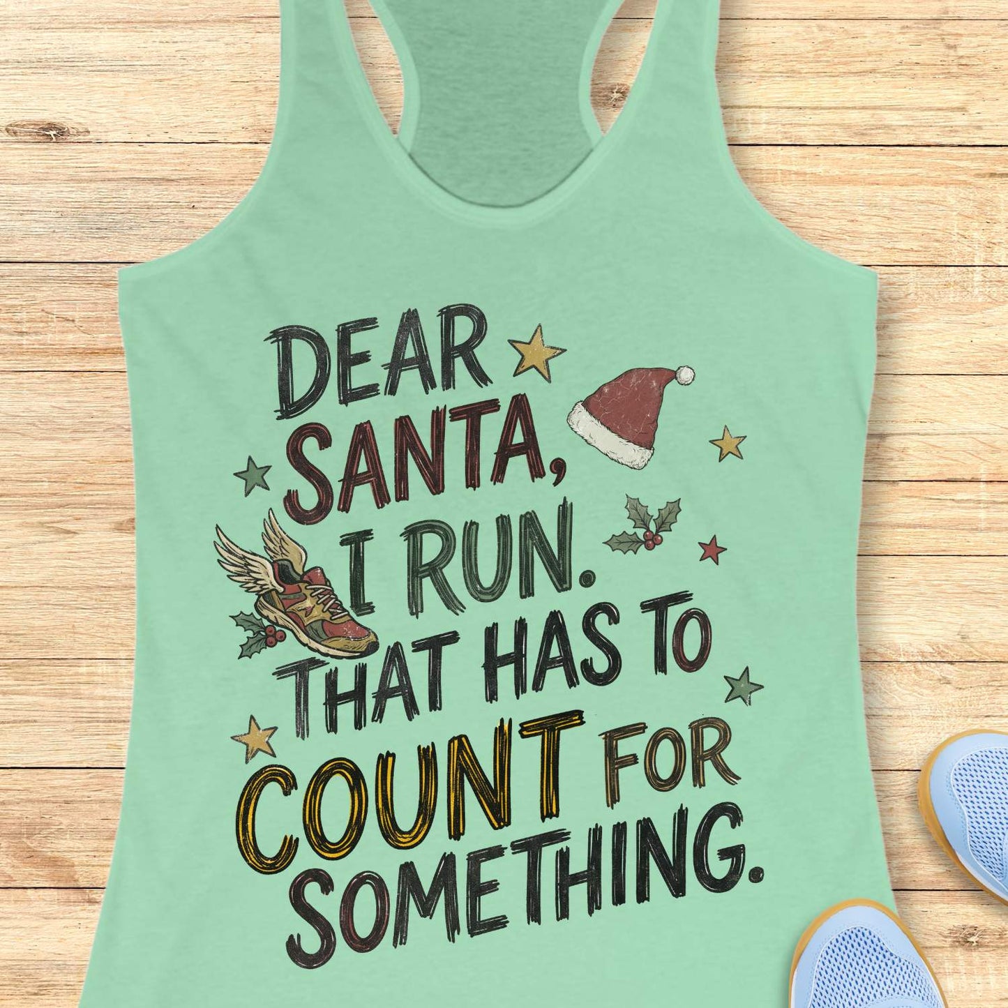 mint racerback tank top displayed on a light wooden surface, featuring a festive running-themed typographic design. The graphic includes the humorous text: “DEAR SANTA, I RUN. THAT HAS TO COUNT FOR SOMETHING.” Decorative holiday elements surround the text, including a red Santa hat, a winged running shoe, holly sprigs with berries, and colorful stars. The word “COUNT” is highlighted in yellow, while the remaining text appears in black. Partial blue athletic shoes are visible in the lower corners, rei