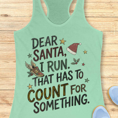 mint racerback tank top displayed on a light wooden surface, featuring a festive running-themed typographic design. The graphic includes the humorous text: “DEAR SANTA, I RUN. THAT HAS TO COUNT FOR SOMETHING.” Decorative holiday elements surround the text, including a red Santa hat, a winged running shoe, holly sprigs with berries, and colorful stars. The word “COUNT” is highlighted in yellow, while the remaining text appears in black. Partial blue athletic shoes are visible in the lower corners, rei