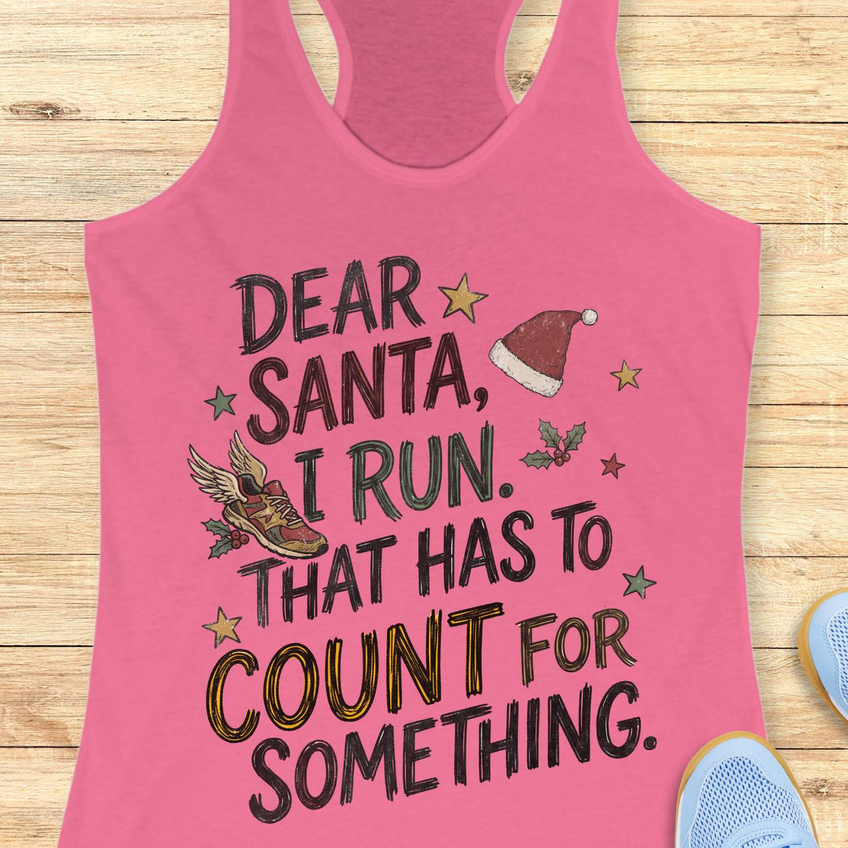 pink racerback tank top displayed on a light wooden surface, featuring a festive running-themed typographic design. The graphic includes the humorous text: “DEAR SANTA, I RUN. THAT HAS TO COUNT FOR SOMETHING.” Decorative holiday elements surround the text, including a red Santa hat, a winged running shoe, holly sprigs with berries, and colorful stars. The word “COUNT” is highlighted in yellow, while the remaining text appears in black. Partial blue athletic shoes are visible in the lower corners, rei