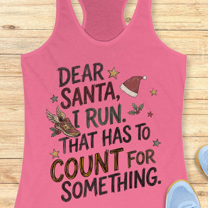 pink racerback tank top displayed on a light wooden surface, featuring a festive running-themed typographic design. The graphic includes the humorous text: “DEAR SANTA, I RUN. THAT HAS TO COUNT FOR SOMETHING.” Decorative holiday elements surround the text, including a red Santa hat, a winged running shoe, holly sprigs with berries, and colorful stars. The word “COUNT” is highlighted in yellow, while the remaining text appears in black. Partial blue athletic shoes are visible in the lower corners, rei