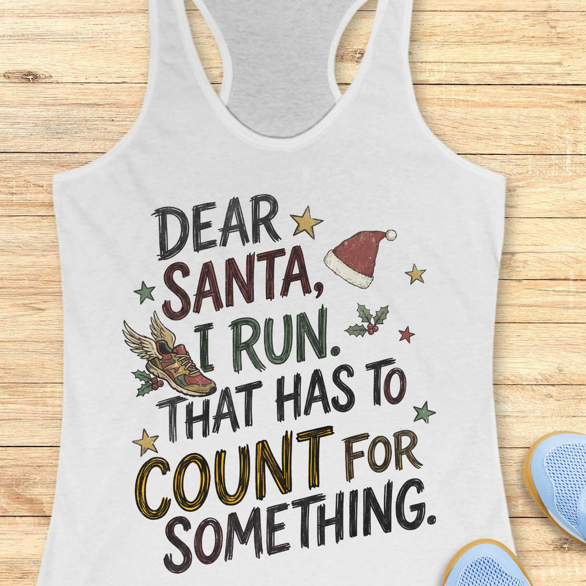 white racerback tank top displayed on a light wooden surface, featuring a festive running-themed typographic design. The graphic includes the humorous text: “DEAR SANTA, I RUN. THAT HAS TO COUNT FOR SOMETHING.” Decorative holiday elements surround the text, including a red Santa hat, a winged running shoe, holly sprigs with berries, and colorful stars. The word “COUNT” is highlighted in yellow, while the remaining text appears in black. Partial blue athletic shoes are visible in the lower corners, rei