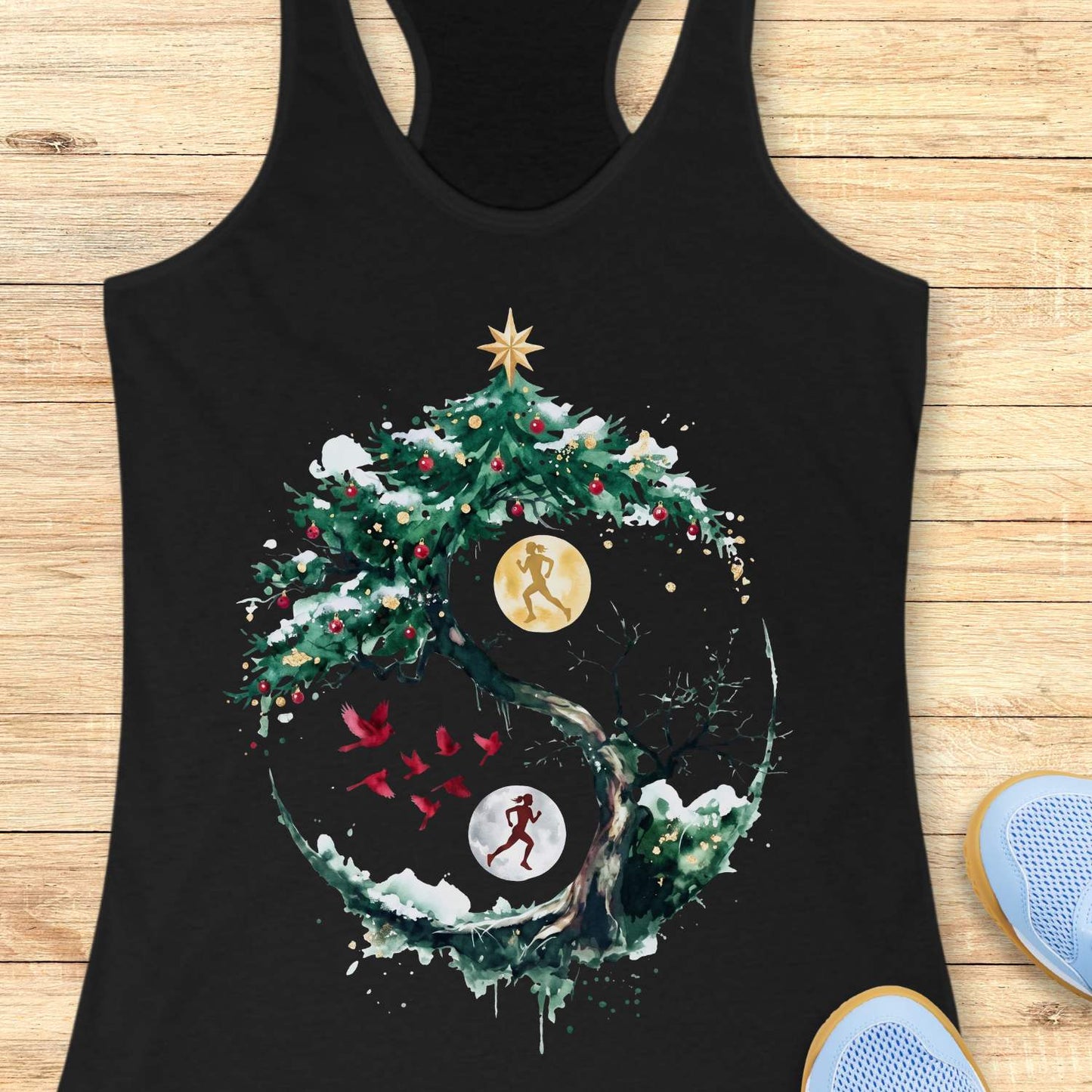 Black racerback tank top displayed on a light wooden surface, featuring an artistic winter-themed running design. The graphic shows a snow-covered, circular tree with evergreen branches decorated with red ornaments and topped with a gold star. Inside the circular composition, two silhouetted runners appear against glowing moon-like circles, one gold and one white, creating a yin-yang-inspired effect. Several red birds fly across the center of the design, while snowy details and watercolor-style splashes add