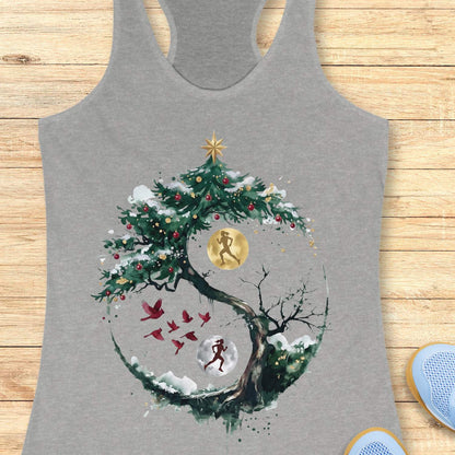 grey racerback tank top displayed on a light wooden surface, featuring an artistic winter-themed running design. The graphic shows a snow-covered, circular tree with evergreen branches decorated with red ornaments and topped with a gold star. Inside the circular composition, two silhouetted runners appear against glowing moon-like circles, one gold and one white, creating a yin-yang-inspired effect. Several red birds fly across the center of the design, while snowy details and watercolor-style splashes add