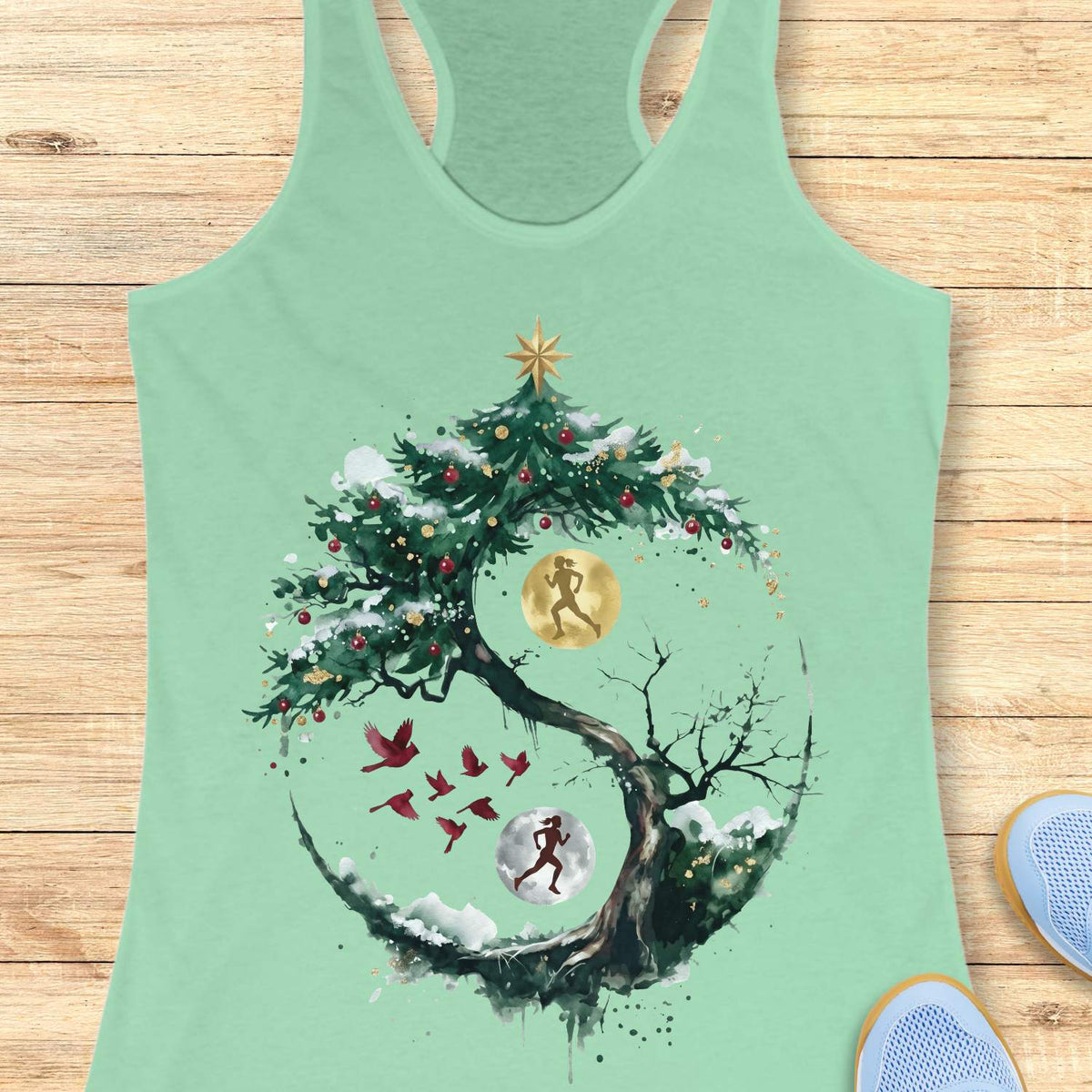 mint racerback tank top displayed on a light wooden surface, featuring an artistic winter-themed running design. The graphic shows a snow-covered, circular tree with evergreen branches decorated with red ornaments and topped with a gold star. Inside the circular composition, two silhouetted runners appear against glowing moon-like circles, one gold and one white, creating a yin-yang-inspired effect. Several red birds fly across the center of the design, while snowy details and watercolor-style splashes add