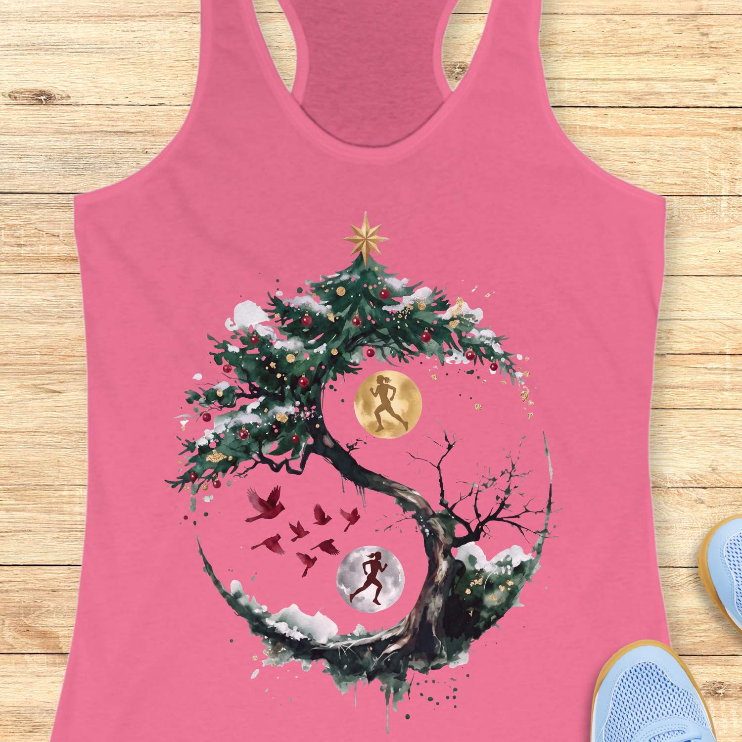 pink racerback tank top displayed on a light wooden surface, featuring an artistic winter-themed running design. The graphic shows a snow-covered, circular tree with evergreen branches decorated with red ornaments and topped with a gold star. Inside the circular composition, two silhouetted runners appear against glowing moon-like circles, one gold and one white, creating a yin-yang-inspired effect. Several red birds fly across the center of the design, while snowy details and watercolor-style splashes add