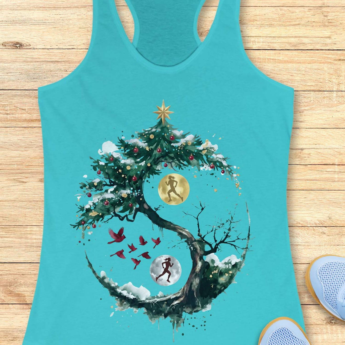 blue racerback tank top displayed on a light wooden surface, featuring an artistic winter-themed running design. The graphic shows a snow-covered, circular tree with evergreen branches decorated with red ornaments and topped with a gold star. Inside the circular composition, two silhouetted runners appear against glowing moon-like circles, one gold and one white, creating a yin-yang-inspired effect. Several red birds fly across the center of the design, while snowy details and watercolor-style splashes add