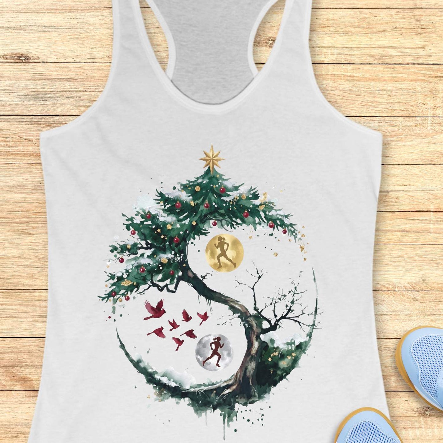 white racerback tank top displayed on a light wooden surface, featuring an artistic winter-themed running design. The graphic shows a snow-covered, circular tree with evergreen branches decorated with red ornaments and topped with a gold star. Inside the circular composition, two silhouetted runners appear against glowing moon-like circles, one gold and one white, creating a yin-yang-inspired effect. Several red birds fly across the center of the design, while snowy details and watercolor-style splashes add