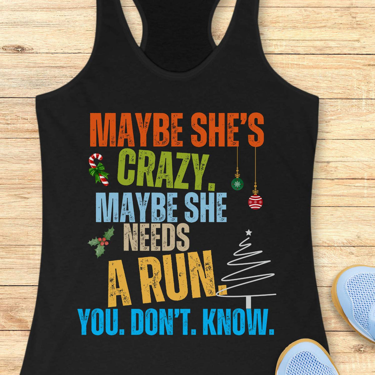 Black racerback tank top displayed on a light wooden surface, featuring a humorous Christmas-themed running design with bold, multicolored typography. The text reads: “MAYBE SHE’S CRAZY. MAYBE SHE NEEDS A RUN. YOU. DON’T. KNOW.” Decorative holiday elements include a candy cane with a green bow, holly sprigs with red berries, hanging Christmas ornaments, and a minimalist white Christmas tree graphic. The design uses vibrant orange, green, blue, yellow, and beige lettering that stands out against the black fa