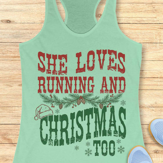 Mint green racerback tank top featuring a distressed vintage-style Christmas running design. The text reads, “She Loves Running and Christmas Too” in red and green lettering. Evergreen pine branches with pinecones and berries separate the lines of text, while a Santa hat rests on the word “Christmas.” Decorative snowflakes complete the festive holiday-themed graphic.