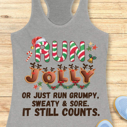 Heather gray racerback tank top featuring a festive running-themed Christmas design. The text reads, “RUN JOLLY or just run grumpy, sweaty & sore. It still counts.” The word “RUN” is styled with red, green, and white candy cane stripes and topped with a Santa hat. The word “JOLLY” is decorated with reindeer antlers, red noses, and holiday greenery with bows. Additional Christmas accents include a candy cane, holly bell, colorful stars, and festive decorations throughout the design.