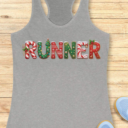 grey racerback tank top featuring the word “RUNNER” in large festive Christmas-themed letters. The letters are decorated with candy cane stripes, holiday sweater patterns, ornaments, holly leaves, bells, snowflakes, and Christmas tree designs in red, green, and white. The tank top is displayed on a light wood background with partial blue running shoes visible in the corners.