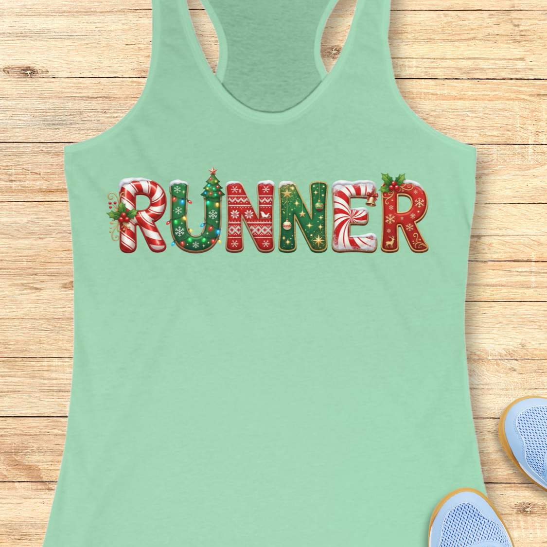 A mint green racerback tank top laid flat on a light wood background, featuring the word 'RUNNER' across the chest in festive holiday-themed lettering with candy canes, Christmas trees, and snowflakes. Partial blue sneakers are visible at the bottom right as styling props.