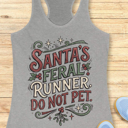 grey racerback tank top featuring the festive phrase “Santa’s Feral Runner, Do Not Pet” in large vintage-style lettering. The design includes red, white, and green text accented with holly leaves, red berries, stars, swirls, and mistletoe-inspired elements, creating a playful Christmas theme for runners. The tank top is displayed on a light wood background with partial blue running shoes visible in the corners.