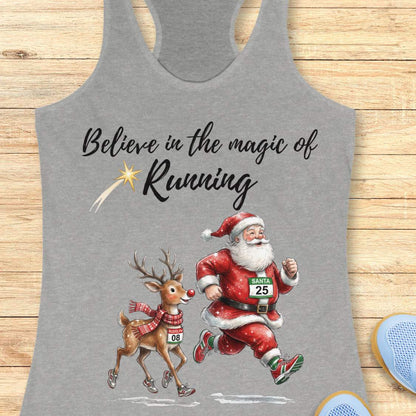 Heather gray racerback tank top featuring the phrase “Believe in the Magic of Running” in black script lettering with a shooting star accent. Below the text, Santa Claus and Rudolph are illustrated running side by side wearing race bibs and running shoes. Santa's bib reads “SANTA 25” and Rudolph's bib reads “RUDOLPH 08.” The festive design combines Christmas and running themes. The tank top is displayed on a light wood background with partial blue running shoes visible in the corners.