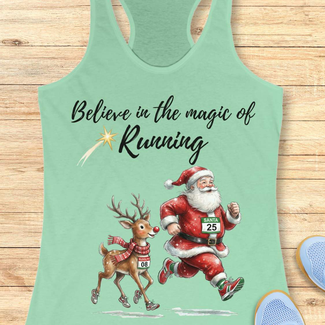 mint racerback tank top featuring the phrase “Believe in the Magic of Running” in black script lettering with a shooting star accent. Below the text, Santa Claus and Rudolph are illustrated running side by side wearing race bibs and running shoes. Santa's bib reads “SANTA 25” and Rudolph's bib reads “RUDOLPH 08.” The festive design combines Christmas and running themes. The tank top is displayed on a light wood background with partial blue running shoes visible in the corners.