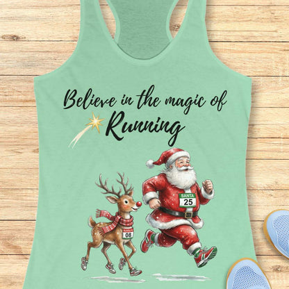 mint racerback tank top featuring the phrase “Believe in the Magic of Running” in black script lettering with a shooting star accent. Below the text, Santa Claus and Rudolph are illustrated running side by side wearing race bibs and running shoes. Santa's bib reads “SANTA 25” and Rudolph's bib reads “RUDOLPH 08.” The festive design combines Christmas and running themes. The tank top is displayed on a light wood background with partial blue running shoes visible in the corners.