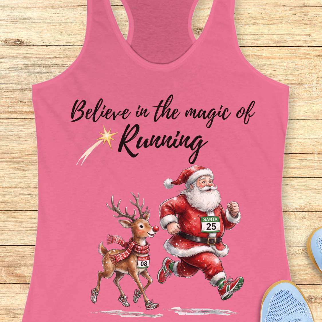 pink racerback tank top featuring the phrase “Believe in the Magic of Running” in black script lettering with a shooting star accent. Below the text, Santa Claus and Rudolph are illustrated running side by side wearing race bibs and running shoes. Santa's bib reads “SANTA 25” and Rudolph's bib reads “RUDOLPH 08.” The festive design combines Christmas and running themes. The tank top is displayed on a light wood background with partial blue running shoes visible in the corners.