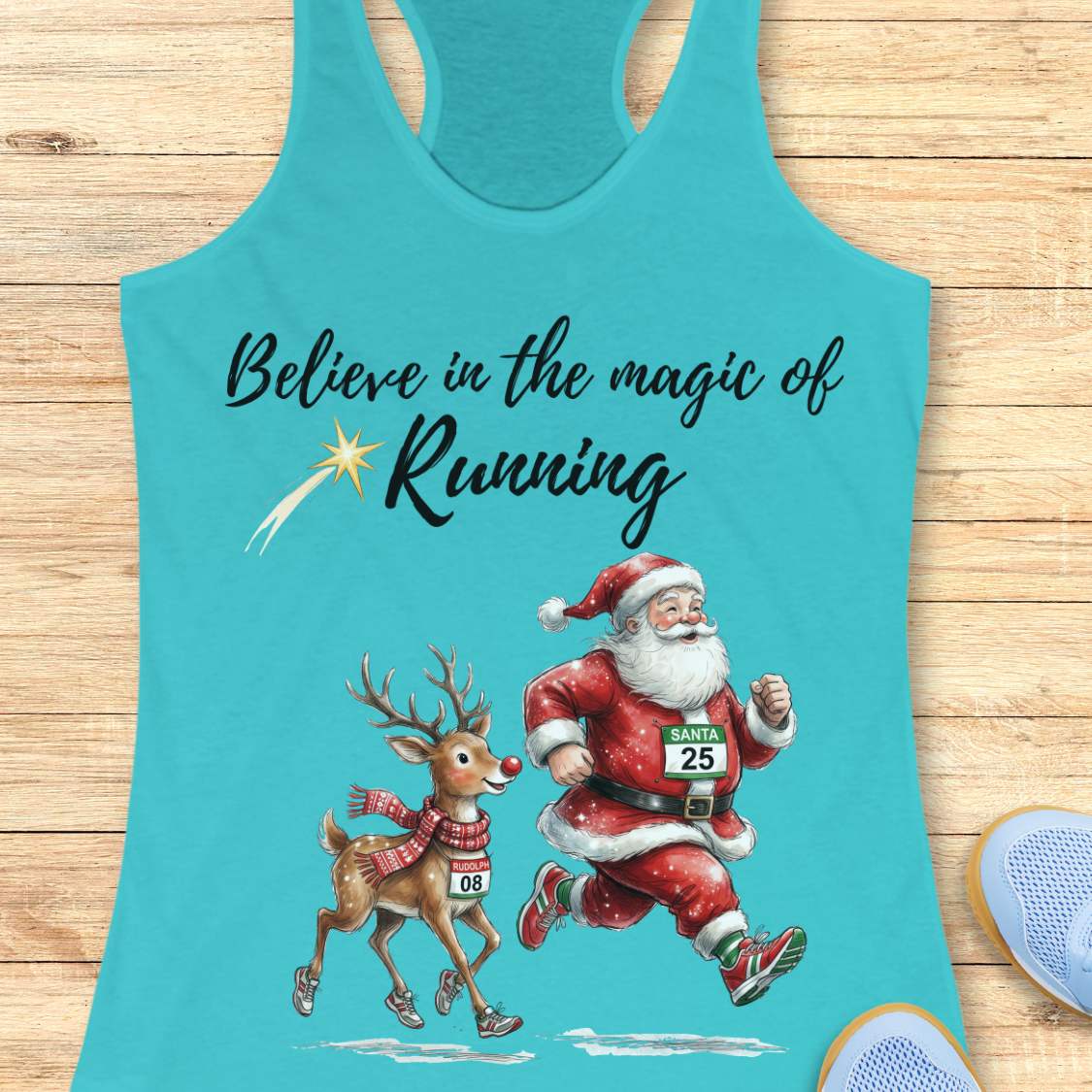 blue racerback tank top featuring the phrase “Believe in the Magic of Running” in black script lettering with a shooting star accent. Below the text, Santa Claus and Rudolph are illustrated running side by side wearing race bibs and running shoes. Santa's bib reads “SANTA 25” and Rudolph's bib reads “RUDOLPH 08.” The festive design combines Christmas and running themes. The tank top is displayed on a light wood background with partial blue running shoes visible in the corners.