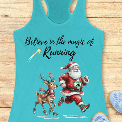 blue racerback tank top featuring the phrase “Believe in the Magic of Running” in black script lettering with a shooting star accent. Below the text, Santa Claus and Rudolph are illustrated running side by side wearing race bibs and running shoes. Santa's bib reads “SANTA 25” and Rudolph's bib reads “RUDOLPH 08.” The festive design combines Christmas and running themes. The tank top is displayed on a light wood background with partial blue running shoes visible in the corners.