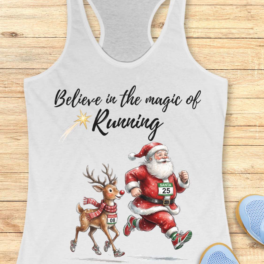 white racerback tank top featuring the phrase “Believe in the Magic of Running” in black script lettering with a shooting star accent. Below the text, Santa Claus and Rudolph are illustrated running side by side wearing race bibs and running shoes. Santa's bib reads “SANTA 25” and Rudolph's bib reads “RUDOLPH 08.” The festive design combines Christmas and running themes. The tank top is displayed on a light wood background with partial blue running shoes visible in the corners.