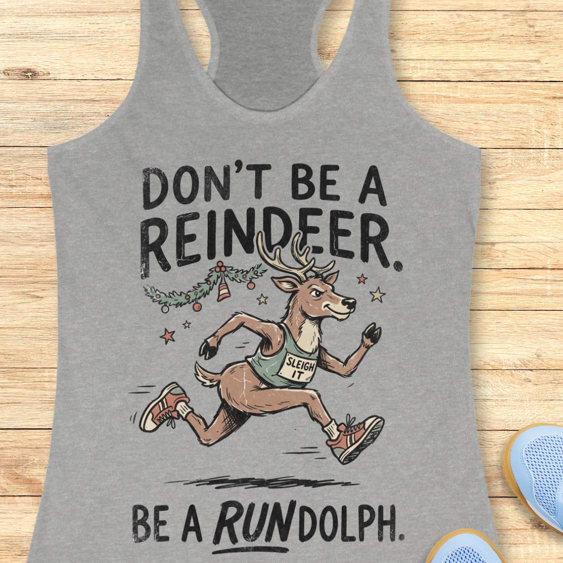 grey racerback tank top featuring the humorous Christmas running phrase “DON’T BE A REINDEER. BE A RUNDOLPH.” in bold black lettering. The design includes a cartoon reindeer wearing running shoes and a race singlet with the words “SLEIGH IT,” sprinting beneath festive garland, ornaments, and stars. The playful holiday-themed artwork combines Christmas cheer with a runner's sense of humor. The tank top is displayed on a light wood background with partial blue running shoes visible in the corners.