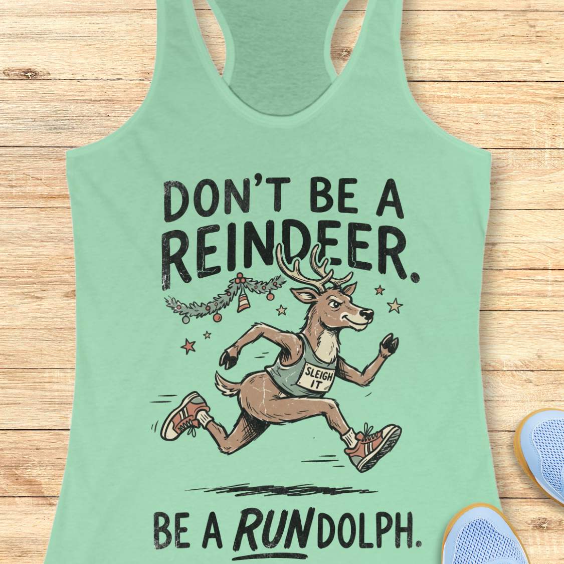 Mint green racerback tank top featuring the humorous Christmas running phrase “DON’T BE A REINDEER. BE A RUNDOLPH.” in bold black lettering. The design includes a cartoon reindeer wearing running shoes and a race singlet with the words “SLEIGH IT,” sprinting beneath festive garland, ornaments, and stars. The playful holiday-themed artwork combines Christmas cheer with a runner's sense of humor. The tank top is displayed on a light wood background with partial blue running shoes visible in the corners.