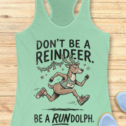Mint green racerback tank top featuring the humorous Christmas running phrase “DON’T BE A REINDEER. BE A RUNDOLPH.” in bold black lettering. The design includes a cartoon reindeer wearing running shoes and a race singlet with the words “SLEIGH IT,” sprinting beneath festive garland, ornaments, and stars. The playful holiday-themed artwork combines Christmas cheer with a runner's sense of humor. The tank top is displayed on a light wood background with partial blue running shoes visible in the corners.