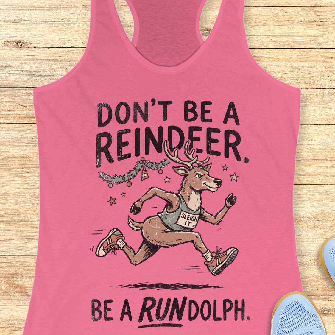 pink racerback tank top featuring the humorous Christmas running phrase “DON’T BE A REINDEER. BE A RUNDOLPH.” in bold black lettering. The design includes a cartoon reindeer wearing running shoes and a race singlet with the words “SLEIGH IT,” sprinting beneath festive garland, ornaments, and stars. The playful holiday-themed artwork combines Christmas cheer with a runner's sense of humor. The tank top is displayed on a light wood background with partial blue running shoes visible in the corners.