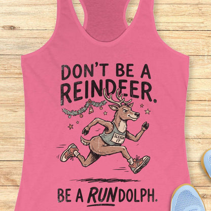 pink racerback tank top featuring the humorous Christmas running phrase “DON’T BE A REINDEER. BE A RUNDOLPH.” in bold black lettering. The design includes a cartoon reindeer wearing running shoes and a race singlet with the words “SLEIGH IT,” sprinting beneath festive garland, ornaments, and stars. The playful holiday-themed artwork combines Christmas cheer with a runner's sense of humor. The tank top is displayed on a light wood background with partial blue running shoes visible in the corners.