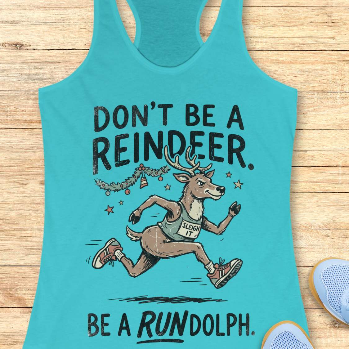 blue racerback tank top featuring the humorous Christmas running phrase “DON’T BE A REINDEER. BE A RUNDOLPH.” in bold black lettering. The design includes a cartoon reindeer wearing running shoes and a race singlet with the words “SLEIGH IT,” sprinting beneath festive garland, ornaments, and stars. The playful holiday-themed artwork combines Christmas cheer with a runner's sense of humor. The tank top is displayed on a light wood background with partial blue running shoes visible in the corners.