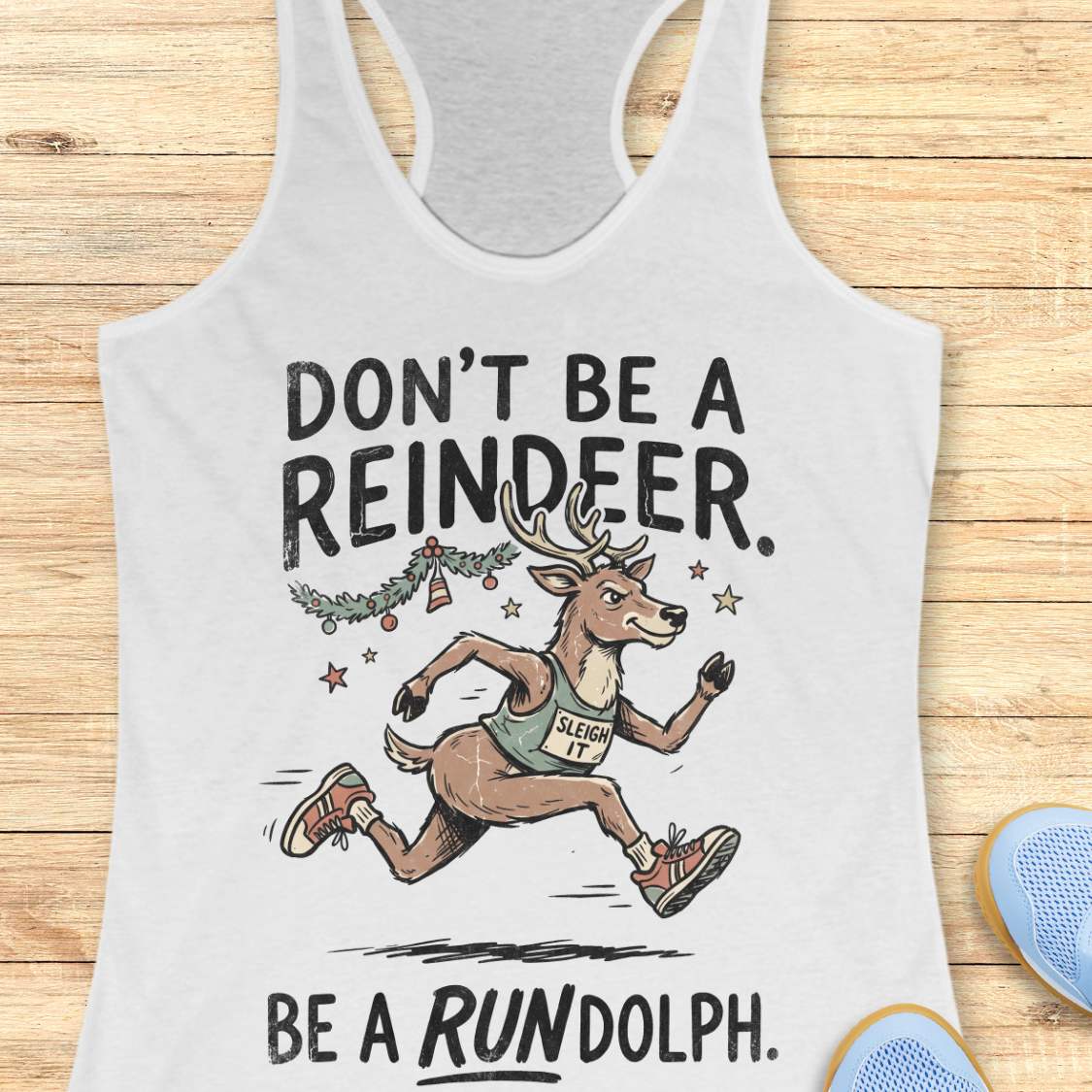 white racerback tank top featuring the humorous Christmas running phrase “DON’T BE A REINDEER. BE A RUNDOLPH.” in bold black lettering. The design includes a cartoon reindeer wearing running shoes and a race singlet with the words “SLEIGH IT,” sprinting beneath festive garland, ornaments, and stars. The playful holiday-themed artwork combines Christmas cheer with a runner's sense of humor. The tank top is displayed on a light wood background with partial blue running shoes visible in the corners.