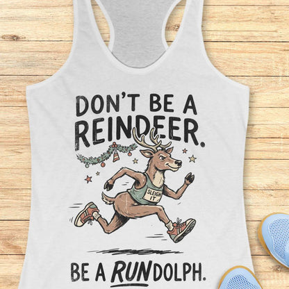 white racerback tank top featuring the humorous Christmas running phrase “DON’T BE A REINDEER. BE A RUNDOLPH.” in bold black lettering. The design includes a cartoon reindeer wearing running shoes and a race singlet with the words “SLEIGH IT,” sprinting beneath festive garland, ornaments, and stars. The playful holiday-themed artwork combines Christmas cheer with a runner's sense of humor. The tank top is displayed on a light wood background with partial blue running shoes visible in the corners.