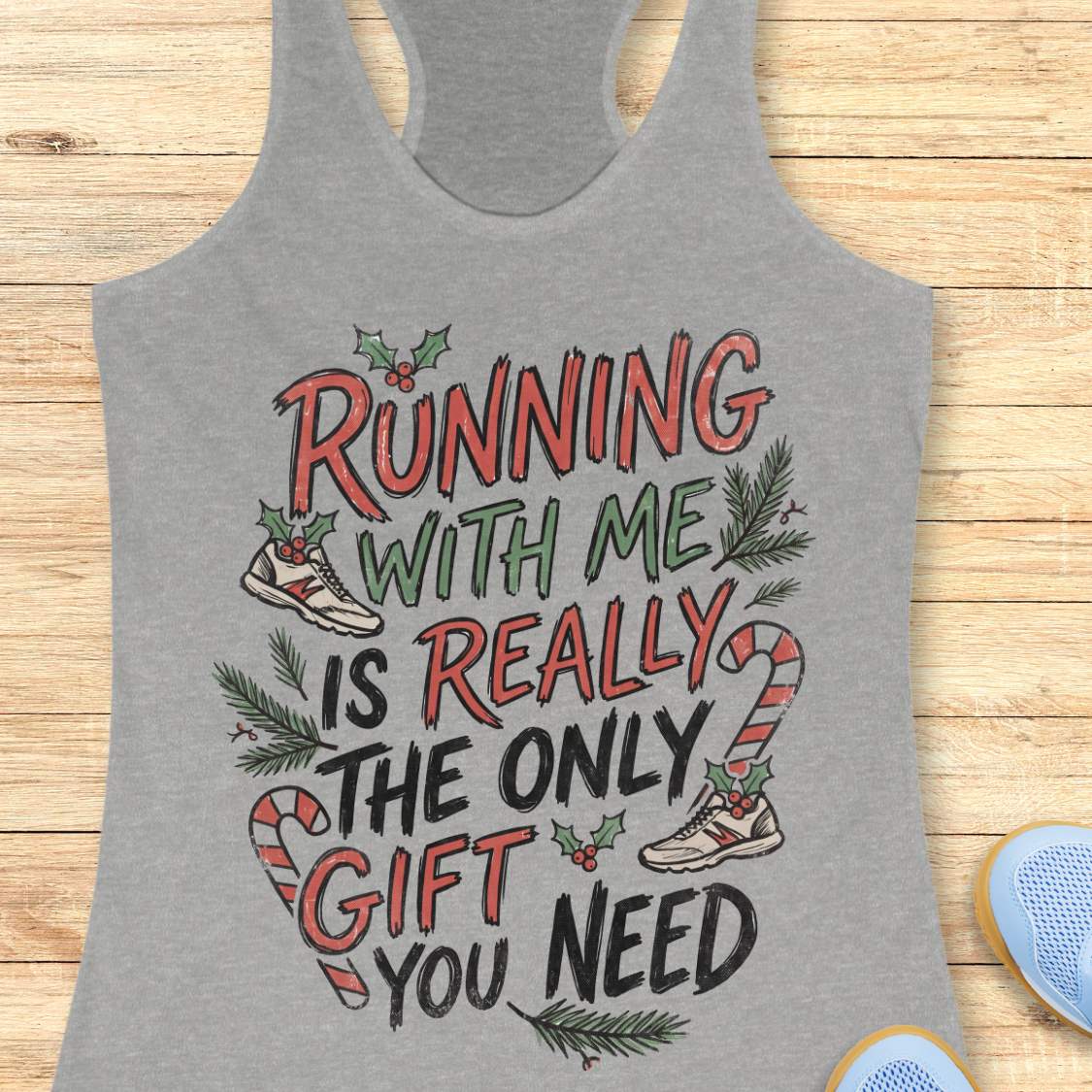 A heather gray racerback tank top with a festive Christmas-themed graphic reading 'RUNNING WITH ME IS REALLY THE ONLY GIFT YOU NEED' in red, green, and black lettering, styled flat on a light wood background.