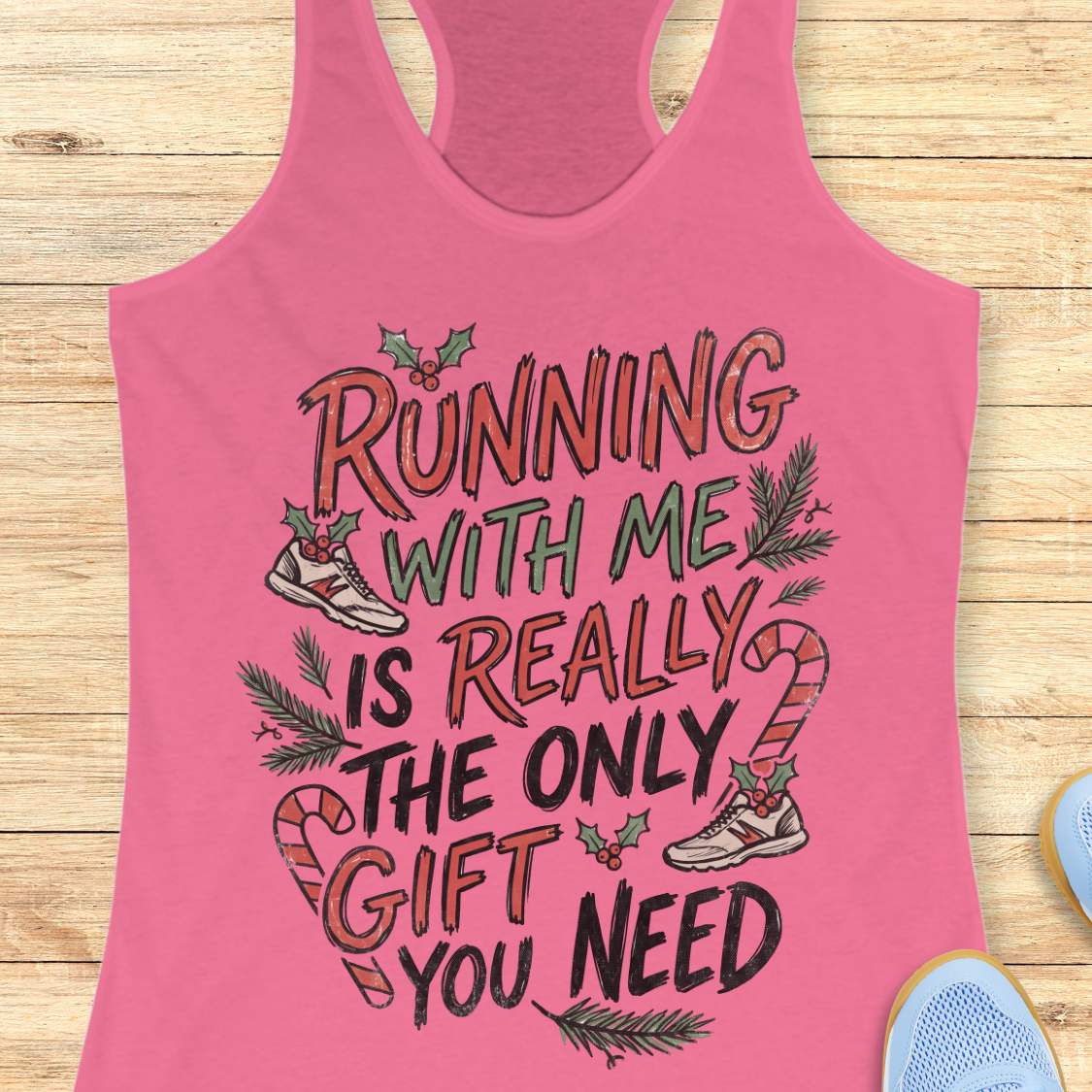 pink racerback tank top featuring the festive running-themed phrase “RUNNING WITH ME IS REALLY THE ONLY GIFT YOU NEED” in large red, green, and black hand-lettered text. The design is decorated with candy canes, holly leaves and berries, evergreen branches, and running shoes accented with holiday greenery. The playful Christmas artwork combines holiday cheer with a runner’s sense of humor. The tank top is displayed on a light wood background with partial blue running shoes visible in the corners.