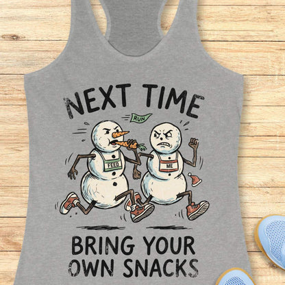 A heather gray racerback tank top laid flat on a light wood background, featuring a cartoon graphic of two snowmen running with the text 'NEXT TIME BRING YOUR OWN SNACKS'.