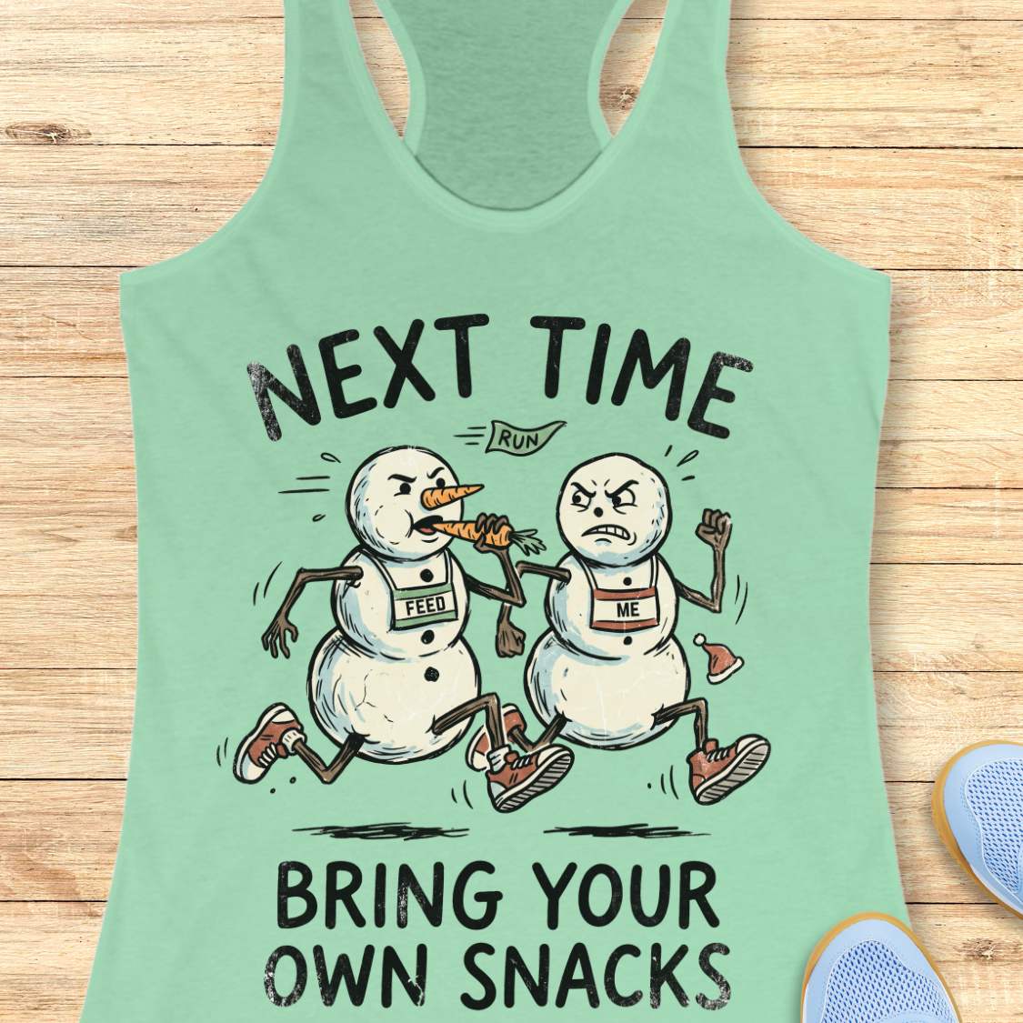 A mint green racerback tank top laid flat on a light wood background, featuring a humorous cartoon graphic of two snowmen running with a carrot. The text reads 'NEXT TIME BRING YOUR OWN SNACKS' with a small 'RUN' banner above the graphic.