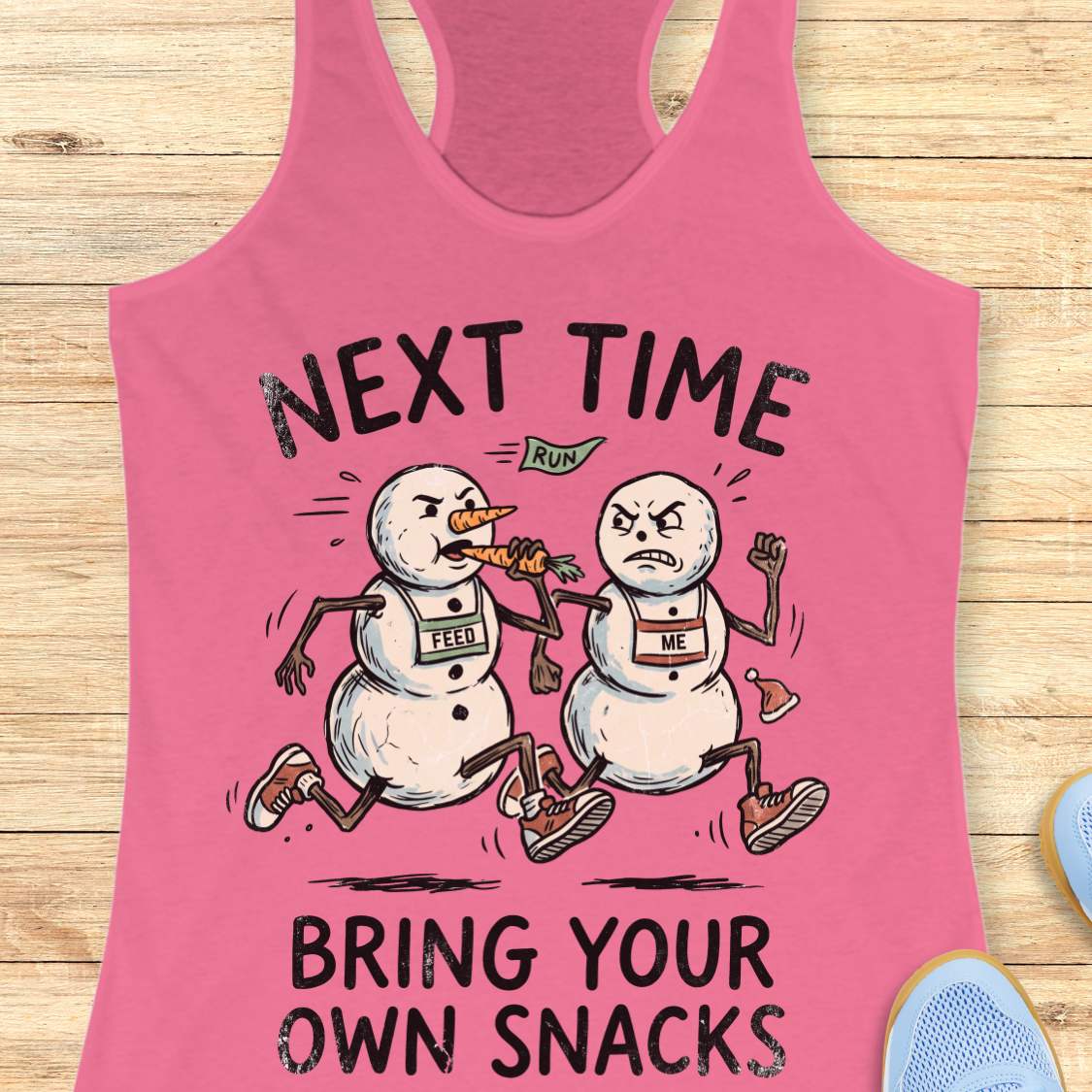 A pink racerback tank top laid flat on a light wood background, featuring a humorous graphic of two cartoon snowmen running with a carrot. The text reads 'NEXT TIME RUN' above the illustration and 'BRING YOUR OWN SNACKS' below it.