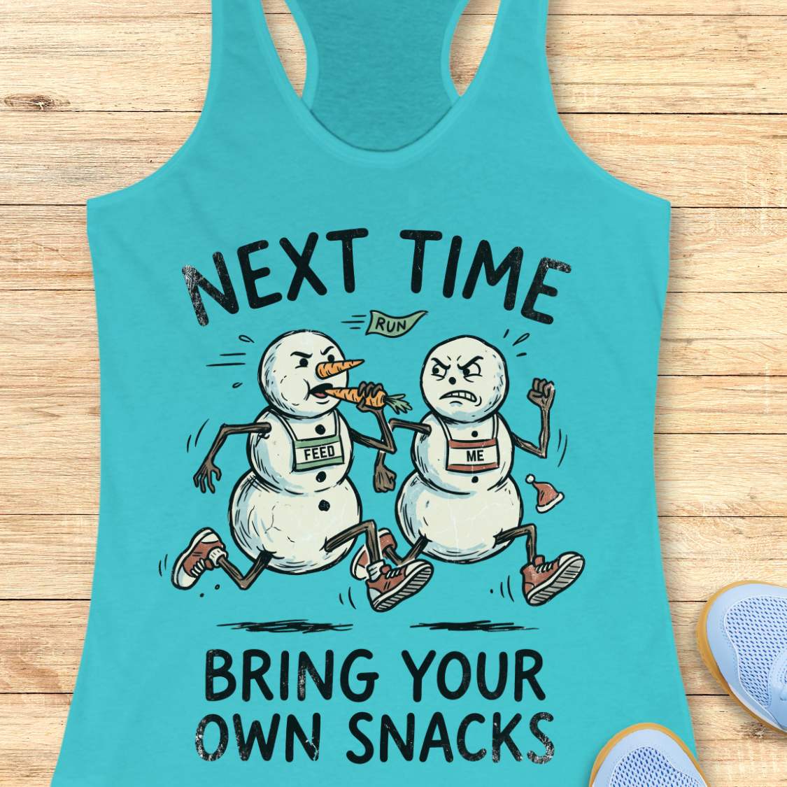 A turquoise racerback tank top laid flat on a light wood surface, featuring a humorous graphic of two snowmen runners with race bibs labeled 'LEAD' and 'ME' under the text 'NEXT TIME BRING YOUR OWN SNACKS'. Partial blue sneakers are visible at the bottom right as styling props.