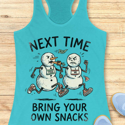 A turquoise racerback tank top laid flat on a light wood surface, featuring a humorous graphic of two snowmen runners with race bibs labeled 'LEAD' and 'ME' under the text 'NEXT TIME BRING YOUR OWN SNACKS'. Partial blue sneakers are visible at the bottom right as styling props.