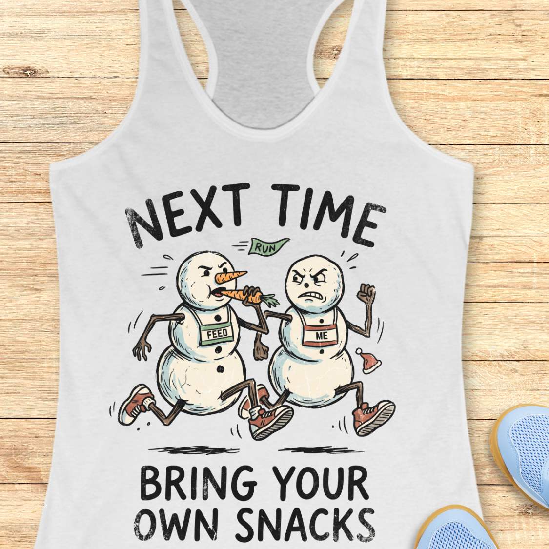 A white racerback tank top laid flat on a light wood background, featuring a cartoon graphic of two snowmen running with snacks and the text 'NEXT TIME BRING YOUR OWN SNACKS'. A blue shoe is partially visible in the bottom right corner.