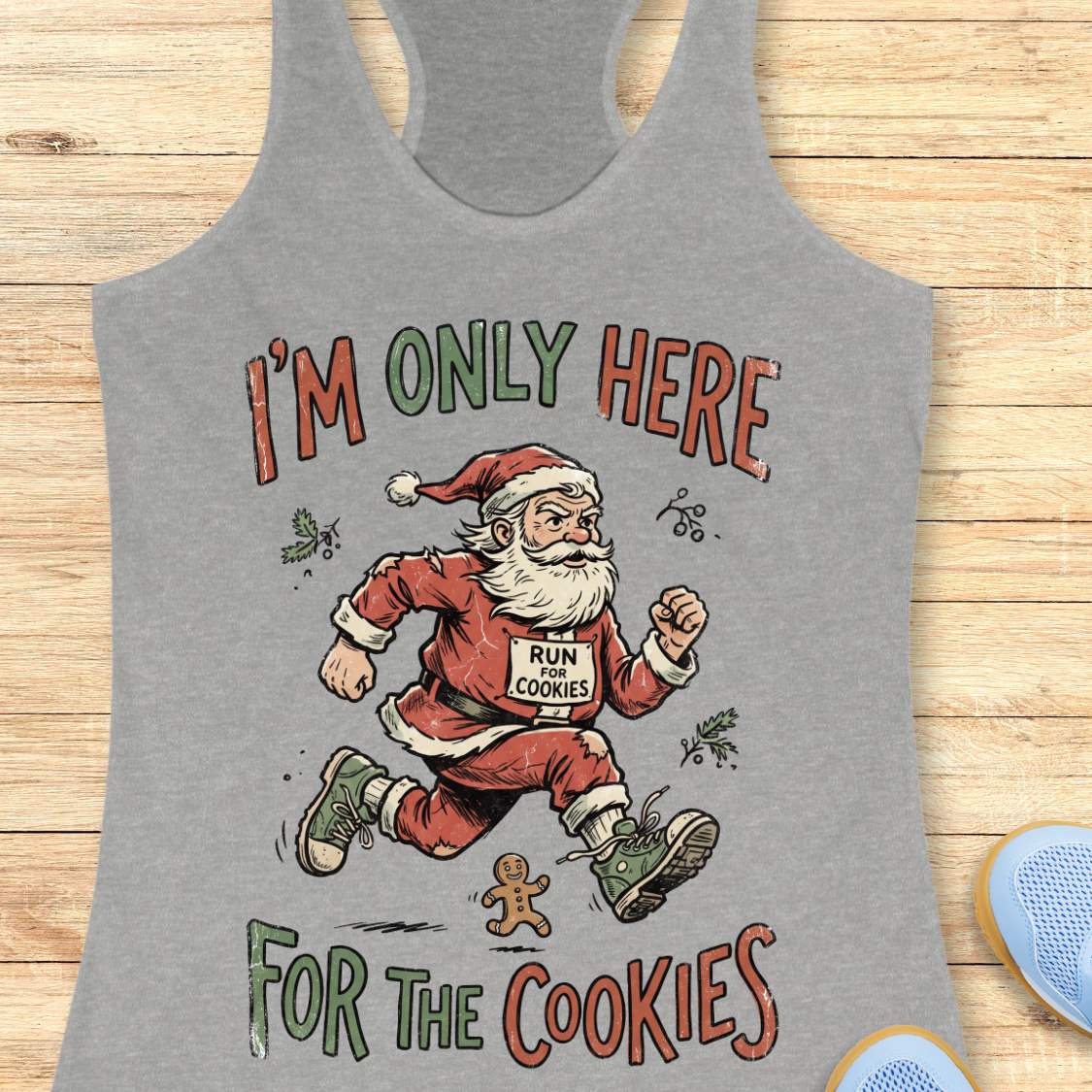 A heather gray racerback tank top featuring a festive graphic of Santa Claus running with a gingerbread cookie on his chest, with the text 'I'M ONLY HERE FOR THE COOKIES' in a holiday font, displayed flat on a light wood background.