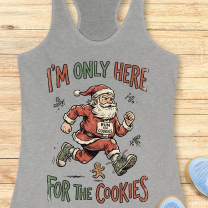 A heather gray racerback tank top featuring a festive graphic of Santa Claus running with a gingerbread cookie on his chest, with the text 'I'M ONLY HERE FOR THE COOKIES' in a holiday font, displayed flat on a light wood background.
