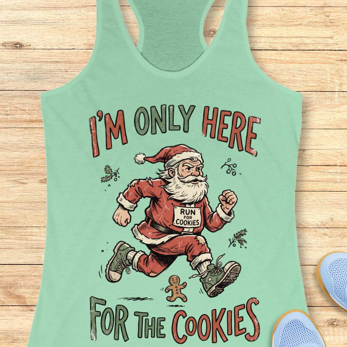A mint green racerback tank top featuring a festive graphic of Santa Claus running with a 'RUN COOKIES' sign, with the text 'I'M ONLY HERE FOR THE COOKIES' in red and dark gray lettering, displayed flat on a light wood background.