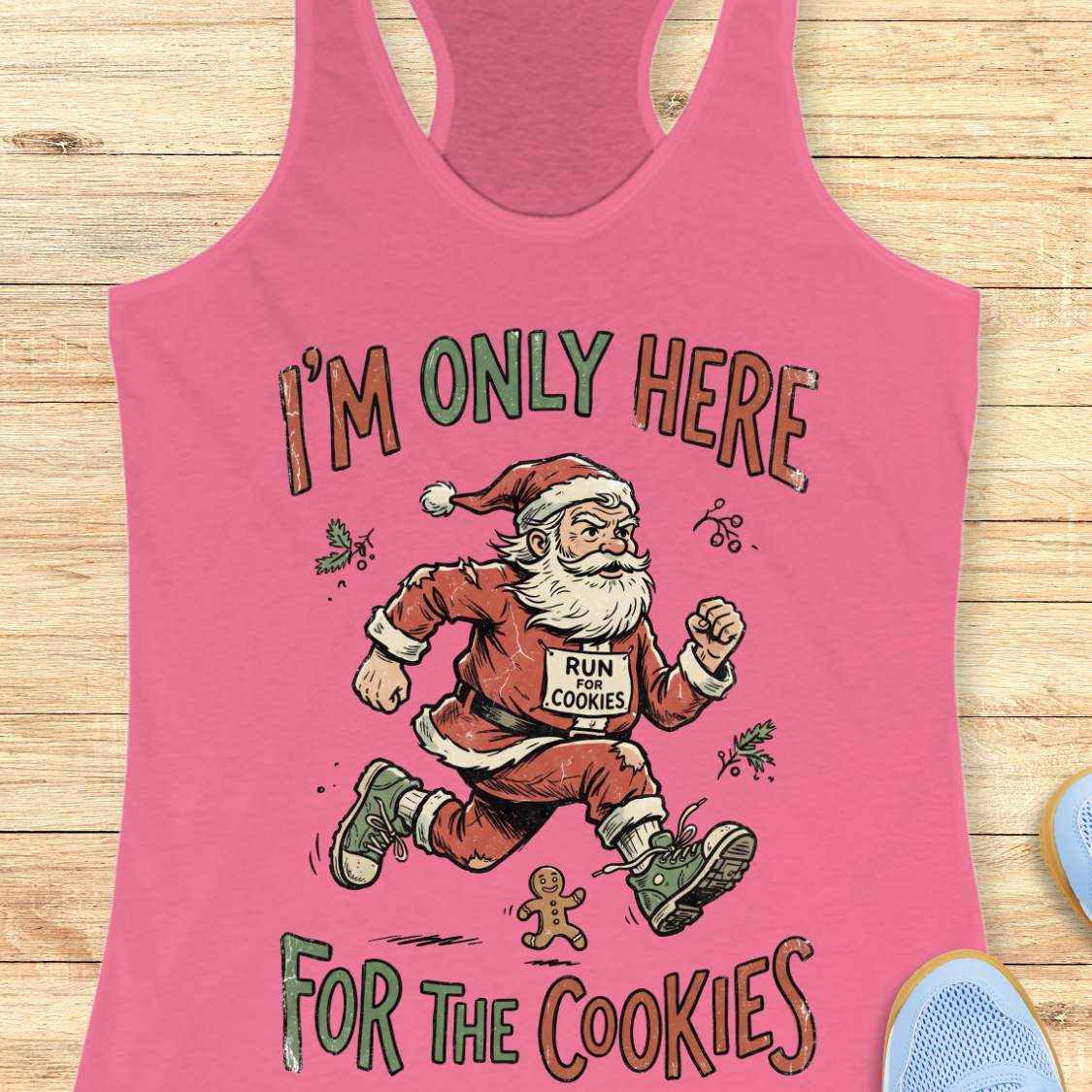 A pink racerback tank top laid flat on a light wood background, featuring a cartoon Santa running with the text 'I'M ONLY HERE FOR THE COOKIES' and a race bib that says 'RUN COOKIES'.