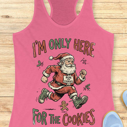 A pink racerback tank top laid flat on a light wood background, featuring a cartoon Santa running with the text 'I'M ONLY HERE FOR THE COOKIES' and a race bib that says 'RUN COOKIES'.