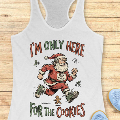 A white racerback tank top laid flat on a light wood background, featuring a colorful graphic of a running Santa Claus with the text 'I'M ONLY HERE FOR THE COOKIES'. Partial blue shoes are visible at the bottom right edge.